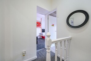 Un couloir avec une rambarde blanche, un miroir à cadre noir et une porte donnant sur une pièce où est accrochée une peinture représentant une fleur rouge.