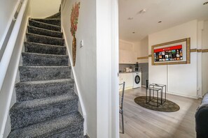 Un escalier mène à un espace de vie comprenant une cuisine, un lave-linge et un écran Netflix fixé au mur.