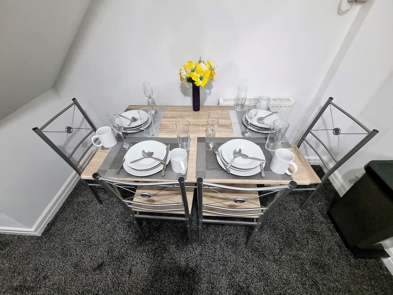 Una mesa de comedor dispuesta con platos, cubiertos y vasos, rodeada de sillas metálicas.