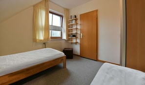 Ein kleiner, ordentlicher Raum mit zwei Einzelbetten, einem Holzschrank, einem Heizkörper und einem Fenster mit Vorhängen.
