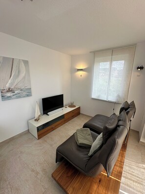 A modern living room with a flat-screen TV, a sofa, a coffee table, a painting, and a window with blinds. - Your dream beachfront vacation rental on Wangerooge (Wangerooge)