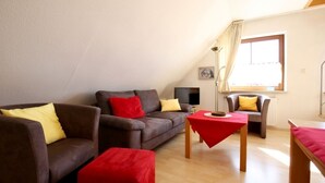 Ein modernes Wohnzimmer mit einer braunen Sofagarnitur, einem roten Hocker, einem kleinen Holztisch und einem Fenster mit Vorhängen.