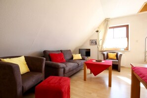 A modern living room with a brown sofa set, a red ottoman, a small wooden table, and a window with curtains. - Pinkenburg Süd in the Kleine Pinkenburg House on Wangerooge (Wangerooge)