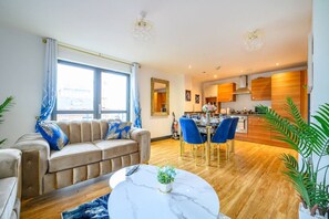 Ein modernes Wohnzimmer mit einer Couch, einem Esstisch und Stühlen, mit einer Küche im Hintergrund und großen Fenstern.