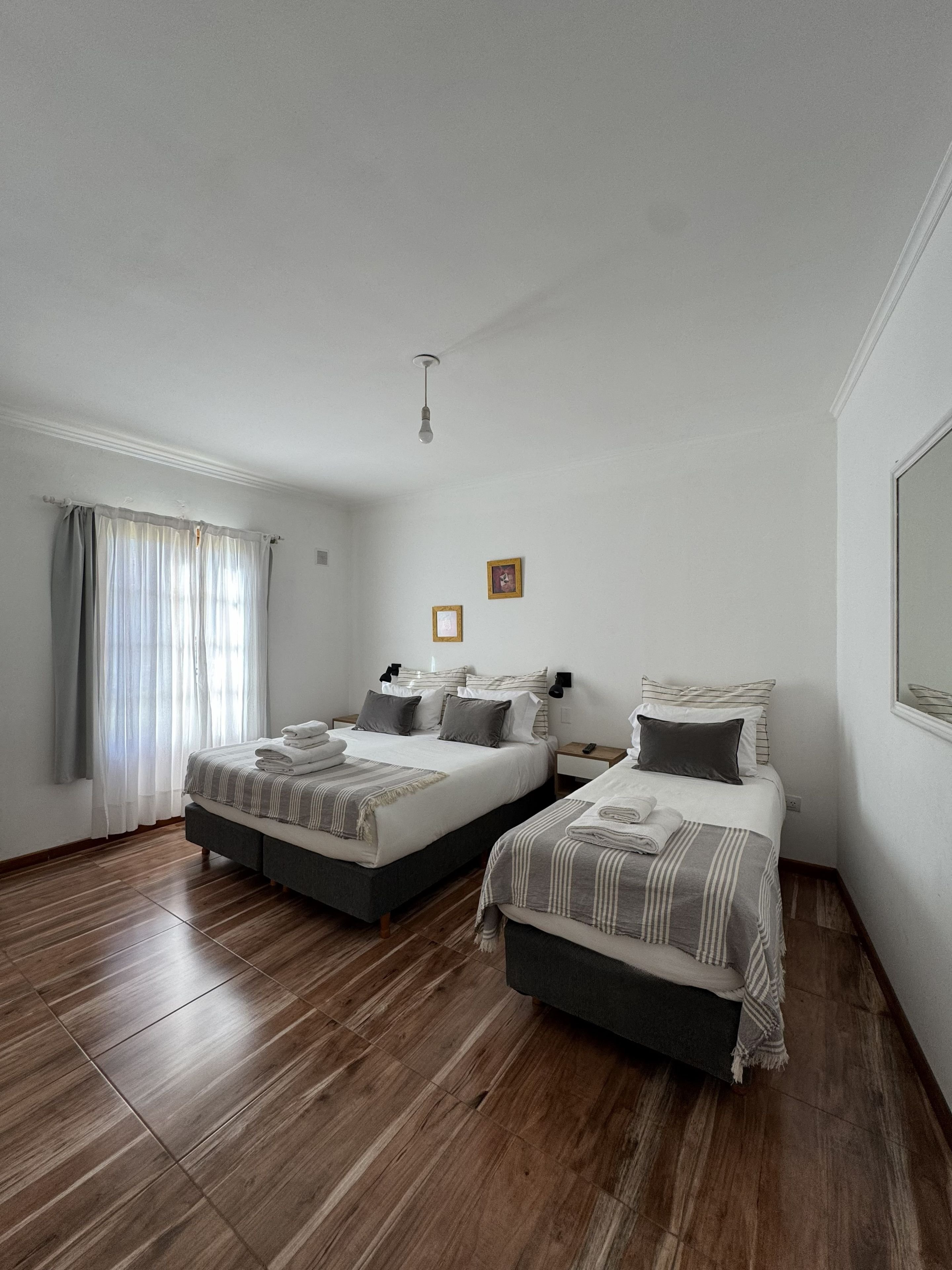 A bedroom with two beds, wooden flooring, a window with curtains, and a single light fixture.