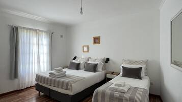A bedroom with two beds, wooden flooring, a window with curtains, and a single light fixture.