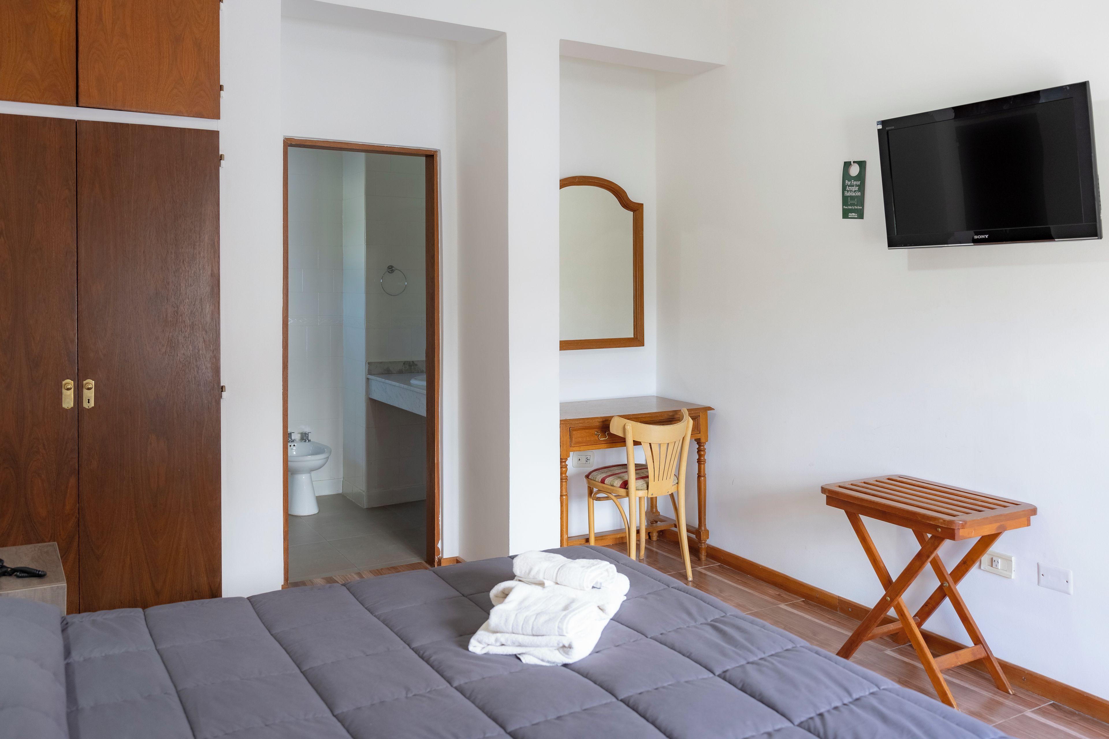 A bedroom with a bed, a wooden desk, a chair, a TV, and a bathroom visible through an open door.