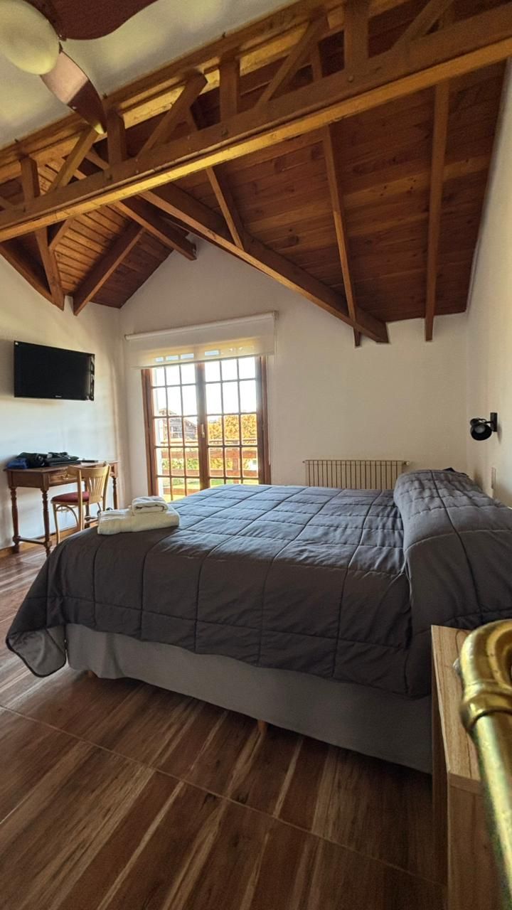 A bedroom with a large bed, wooden ceiling beams, a desk, and a window.