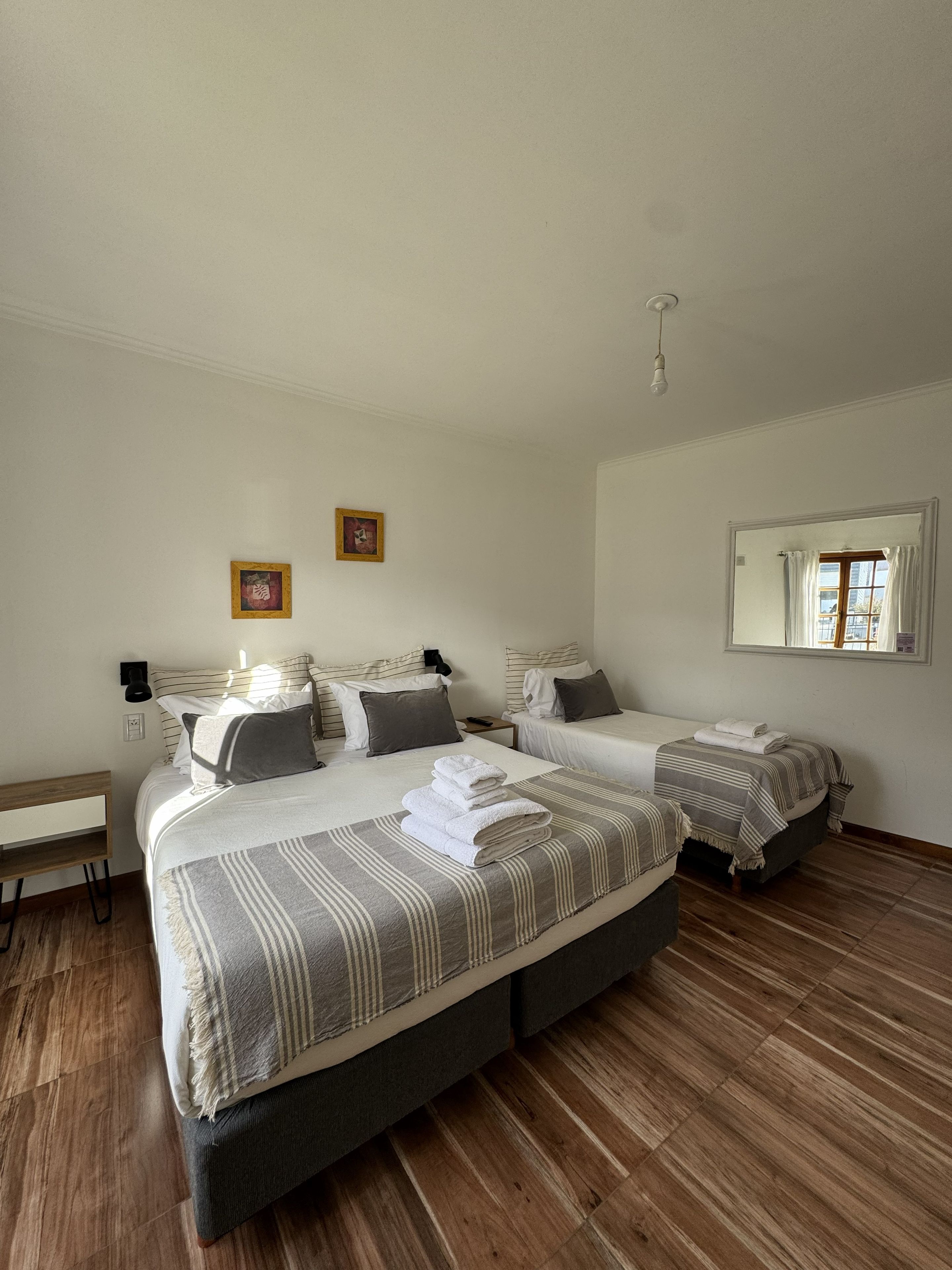 A hotel room with two beds, a bench, a bedside table, a mirror, and a window with curtains.
