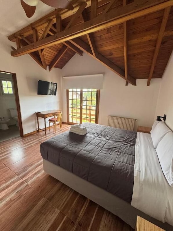 Une chambre avec un grand lit, des poutres apparentes en bois au plafond, une fenêtre, un bureau et un radiateur.