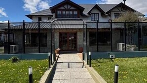 A building with a paved walkway leading to a wooden door, surrounded by a metal fence and greenery. - Hosteria Cumbres Blancas (Esquel)