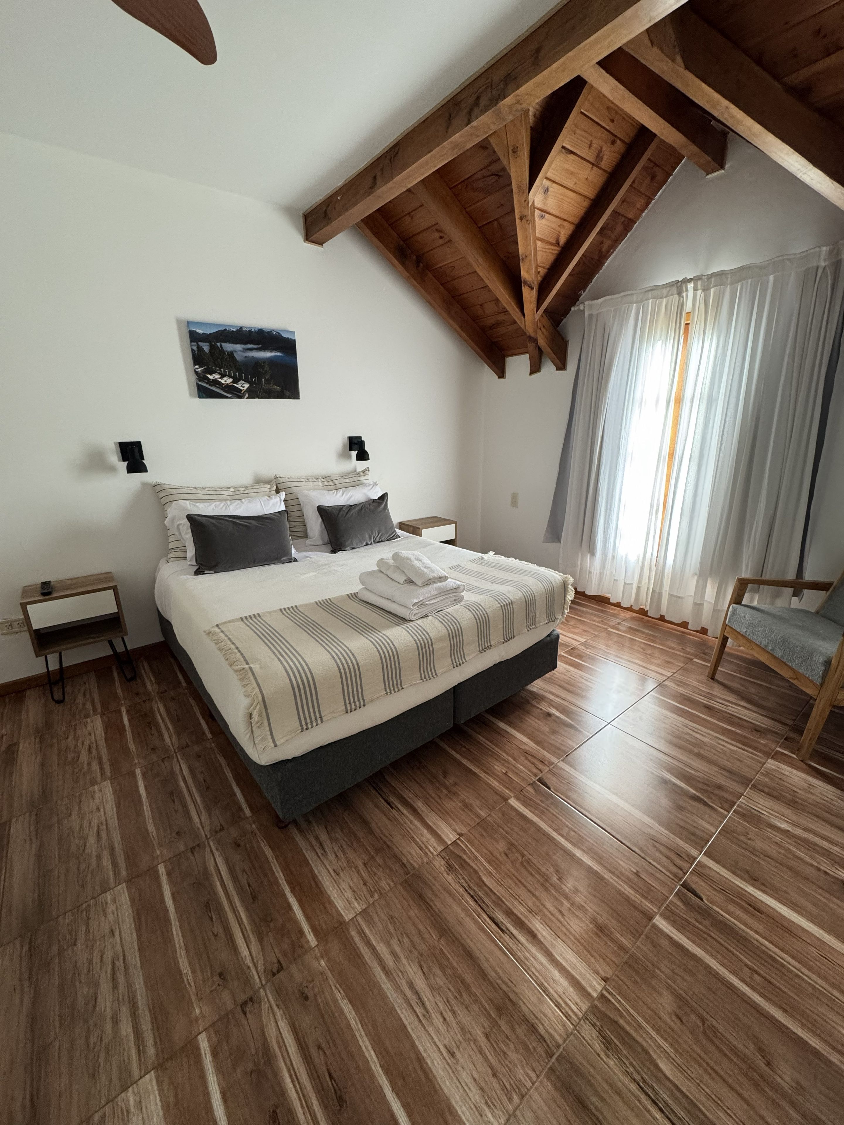 A bedroom with a wooden ceiling, a bed with striped bedding, a nightstand, a chair, and a window with curtains.