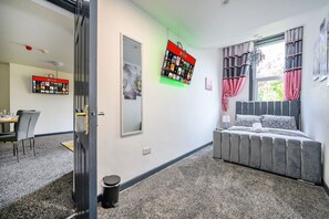 Ett sovrum med en säng, ett fönster med gardiner, en väggmonterad TV och en matplats med stolar och ett bord.