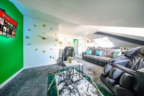 Uma sala de estar moderna com um relógio de parede grande, uma parede verde decorativa, uma poltrona de couro, um sofá com almofadas azul-esverdeadas e uma pequena mesa com uma planta.