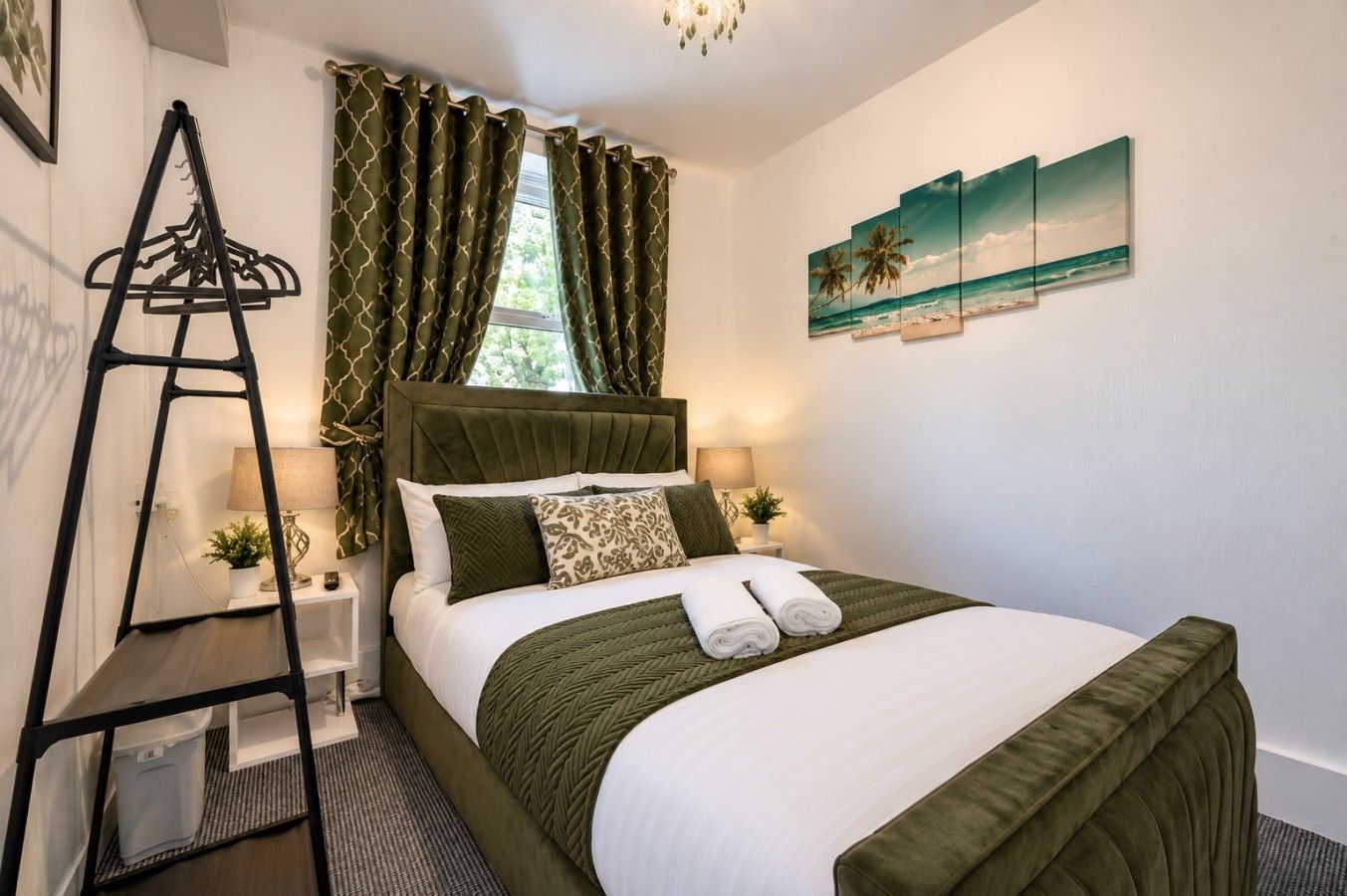 A neatly made bed with a green bedspread and white linens, a bedside table with a lamp, and a window with green curtains.