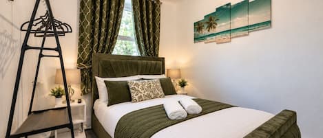 A neatly made bed with a green bedspread and white linens, a bedside table with a lamp, and a window with green curtains.