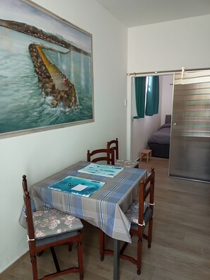 Zona pranzo con tavolo apparecchiato per quattro persone, un quadro raffigurante una veduta costiera e una porta scorrevole che dà su una camera da letto.