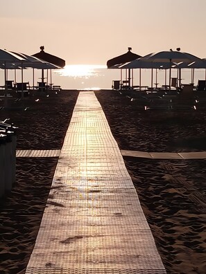 A beach walkway leading to beach umbrellas, with a clear sky and sun setting in the background.