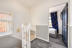 A hallway with a white railing, a window with blinds, and a door leading to a bedroom with a bed and curtains. - Stylish Family Home with Hot Tub (West Yorkshire)