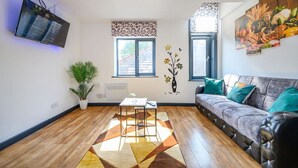 Een woonkamer met een bank, een aan de muur bevestigde televisie, een potplant, een tafel met een vaas en een raam met behang.