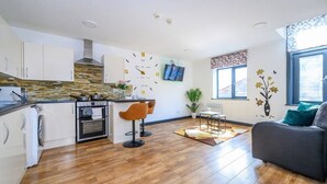 Een moderne keuken met witte kasten, een ingebouwde oven en een kookplaat. Er staan twee barkrukken en een eettafel met een bloemenvaas. De kamer heeft houten vloeren en een groot raam met uitzicht.