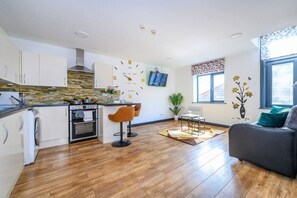 Eine moderne Küche mit weißen Schränken, einem eingebauten Backofen und einem Herd. Zwei Barhocker und ein Esstisch mit einem Blumenstrauß stehen dort. Der Raum hat Holzboden und ein großes Fenster mit Blick.