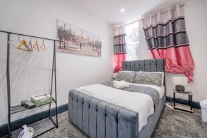 Une chambre à coucher avec un lit, un portant à vêtements, un tableau et une fenêtre avec des rideaux.