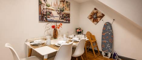 A dining area with a table set for four, white chairs, a vase with flowers, and a painting of a street scene on the wall.