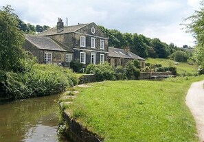 Ein zweistöckiges Steinhaus mit einem Fluss davor, umgeben von viel Grün.