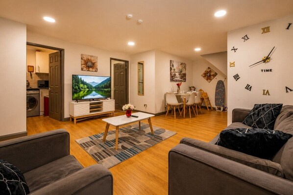 Een moderne woonkamer met een flatscreen televisie, een eethoek met stoelen, een bank en een wandklok.