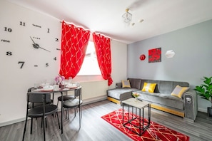 A modern living room with a dining area, a grey sofa with cushions, a red patterned curtain, and a wall clock.