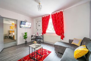 Una sala de estar moderna con un sofá gris, cortinas rojas, un televisor, una mesa de comedor con sillas y una planta en maceta.