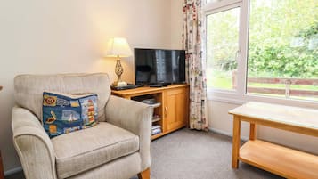 A living room with a beige armchair, a television on a wooden stand, a table, and a window with curtains.