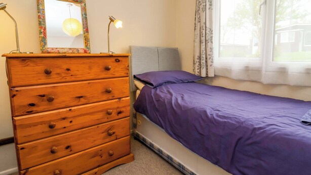 A bedroom with a wooden dresser, a bed with a purple bedspread, a window with curtains, and a wall-mounted mirror. - Sea Breeze (Bideford)