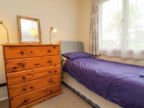 A bedroom with a wooden dresser, a bed with a purple bedspread, a window with curtains, and a wall-mounted mirror. - Sea Breeze (Bideford)