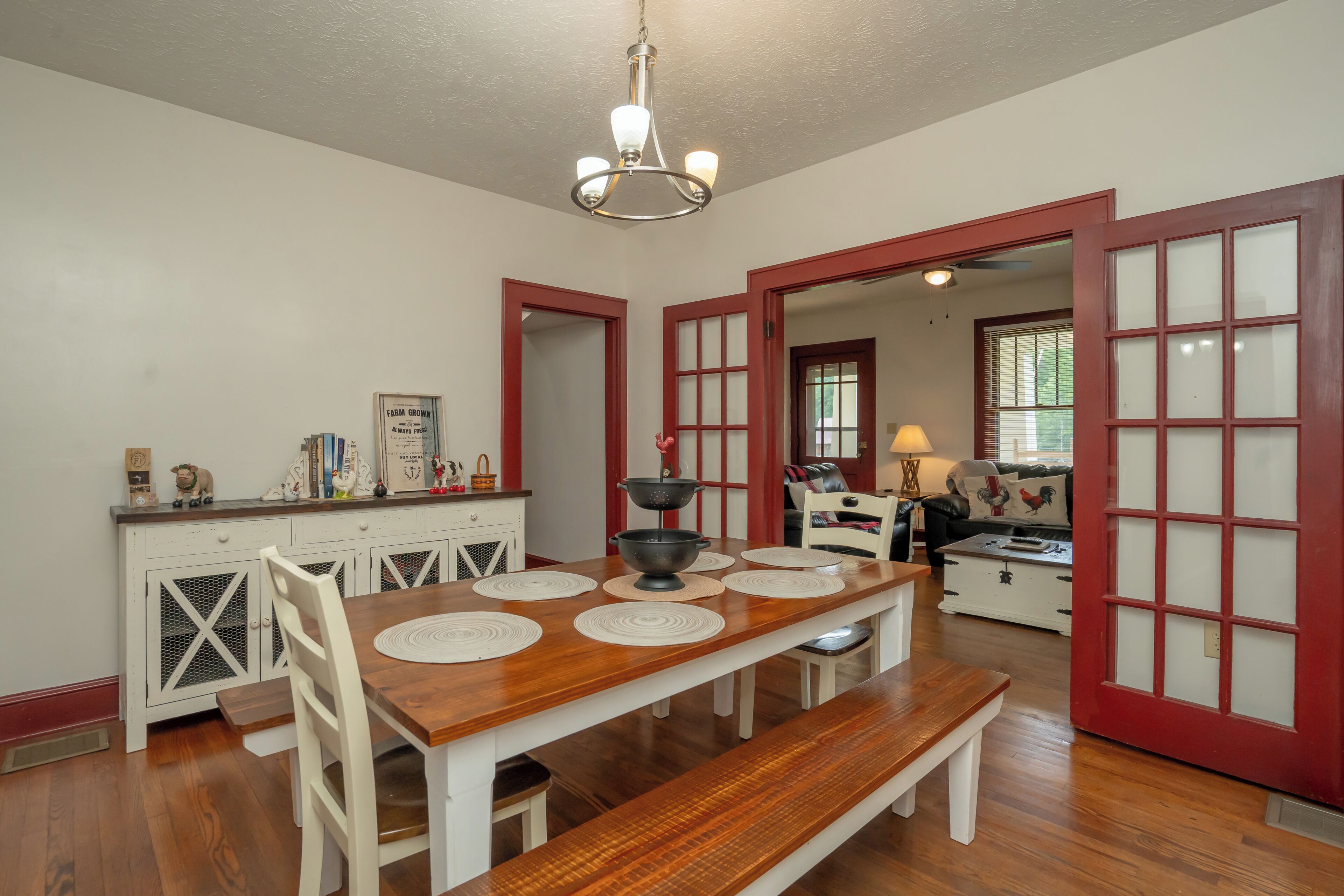 Un coin repas avec une table en bois dressée pour quatre personnes, des chaises blanches et un banc. On y trouve également un coin cuisine avec une porte rouge et un meuble blanc.