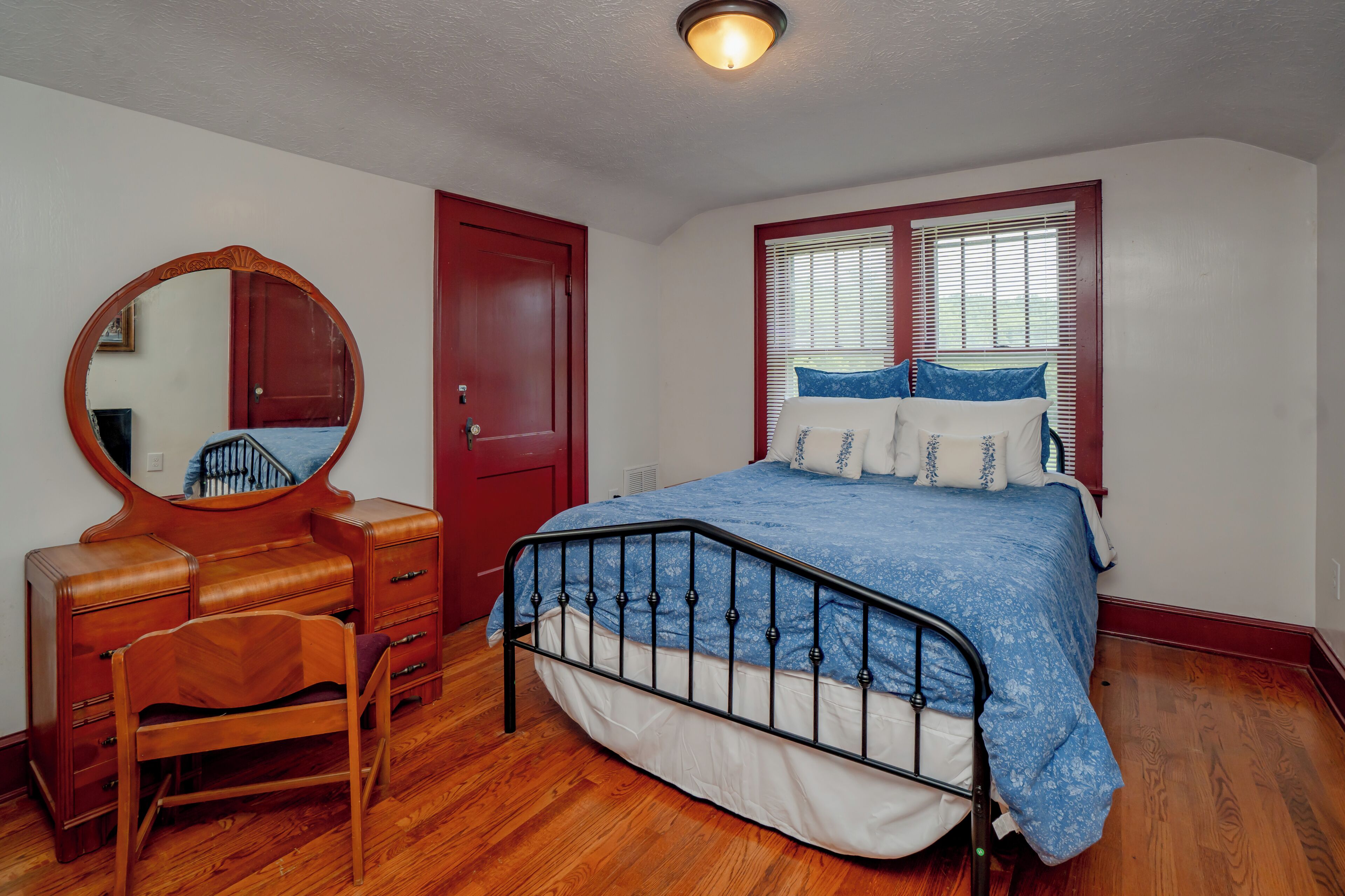 Une chambre avec une commode en bois, un lit recouvert d’une courtepointe à motifs floraux bleus, une porte rouge et une fenêtre avec des stores.