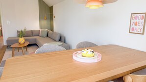 Una sala de estar moderna con una mesa de comedor de madera, un sofá, una pequeña mesita de noche con una planta y un aplique de pared.