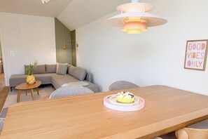 Ein modernes Wohnzimmer mit einem Holzesstisch, einer Couch, einem kleinen Beistelltisch mit Pflanze und einer wandmontierten Leuchte.