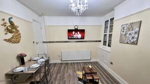 Un coin repas avec une longue table dressée pour quatre personnes, un lustre, un téléviseur et une décoration murale représentant un paon.