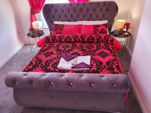 Uma cama com cabeceira estofada, edredom estampado em vermelho e preto, e duas almofadas.