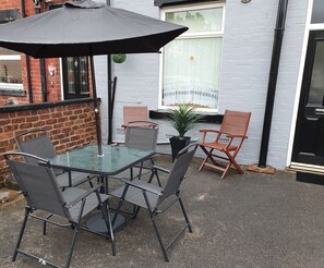 Uma mesa de vidro com cadeiras de metal, um guarda-sol preto e um vaso com planta num pátio.