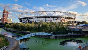 A stadium with a distinctive roof structure, a bridge over a water body, and a large red structure to the left. - Sleep Well - Modern Apartment for 4 near Stations (London)