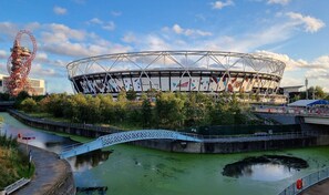 A stadium with a distinctive roof structure, a bridge over a water body, and a large red structure to the left. - Sleep Well - Modern Apartment for 4 near Stations (London)