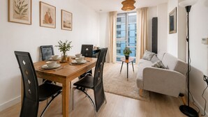 A modern living room with a dining area, featuring a wooden table, black chairs, a grey sofa, and a large window with curtains. - Sleep Well - Modern Apartment for 4 near Stations (London)