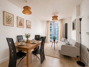 A modern living room with a dining area, featuring a wooden table, black chairs, a grey sofa, and a large window with curtains. - Sleep Well - Modern Apartment for 4 near Stations (London)