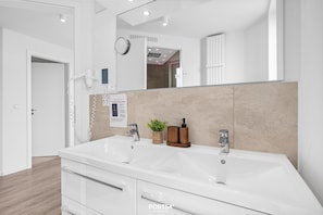 A modern bathroom with a large mirror, two sinks, a towel rack, and a wall-mounted clock. - Küstenbrise holiday flat in Marina Wendtorf with sea views & 159 m² for up to 12 (Wendtorf)