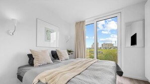 Een slaapkamer met een groot bed, uitzicht op gebouwen door een raam, een aan de muur bevestigde televisie en een lijst met een afbeelding.