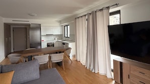 A modern living room with a grey sofa, wooden flooring, a TV, and a kitchen in the background. - Alimos Urban Retreat (Alimos)