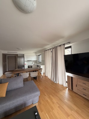 Uma sala de estar moderna com um sofá cinzento, soalho de madeira, uma televisão e uma cozinha ao fundo.